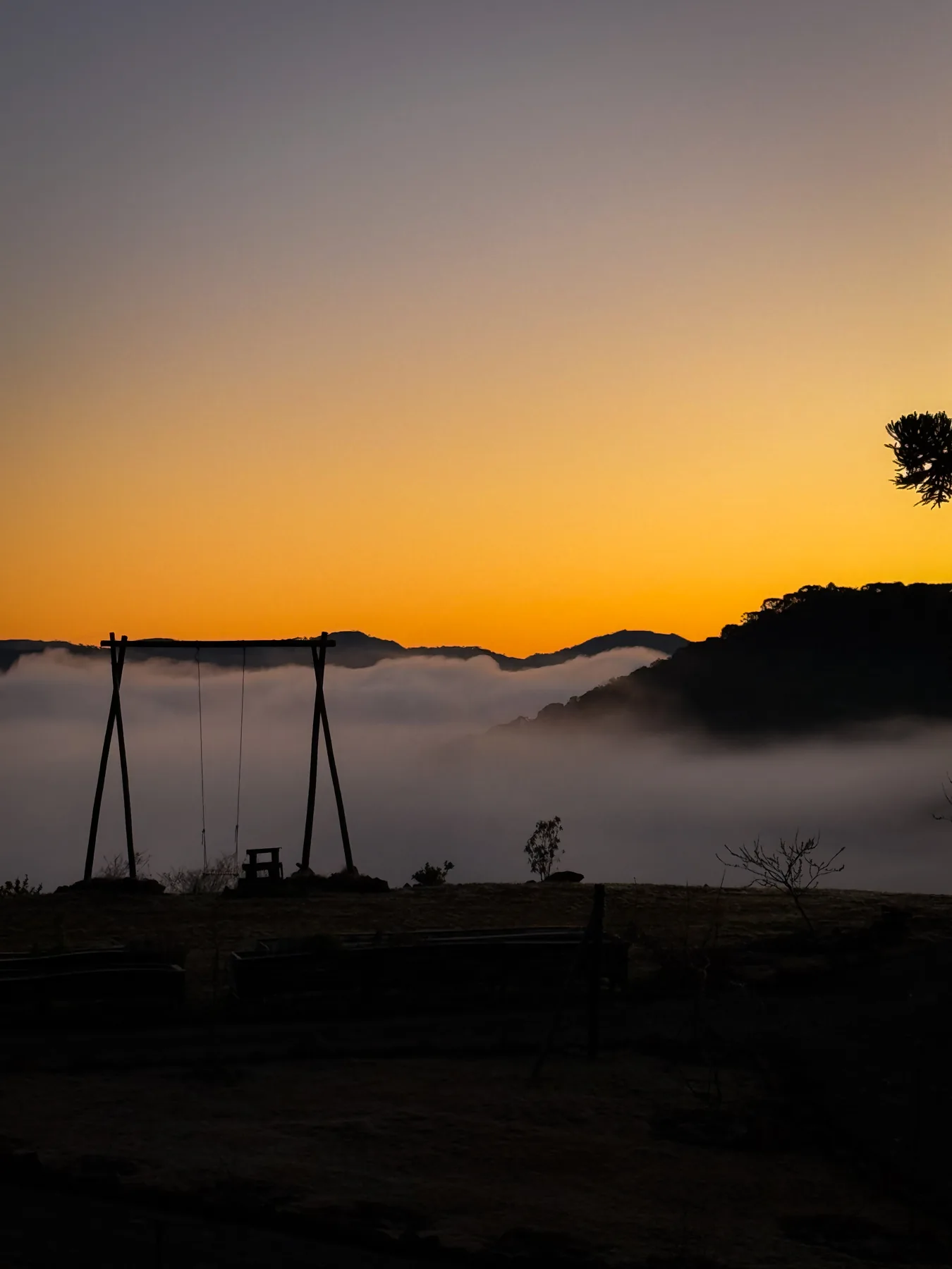 Balanço no alto do morro, recortado contra o céu laranja do amanhecer e a névoa no vale